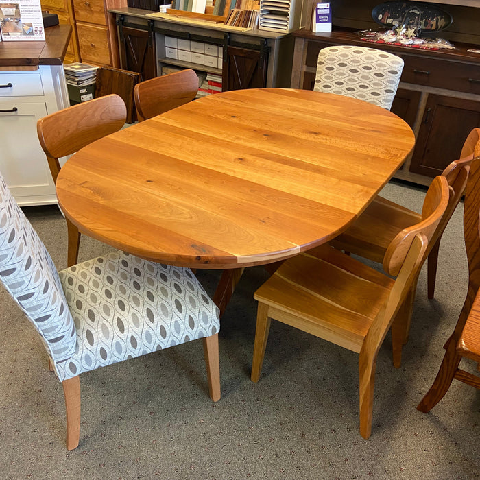 An oval wooden dining table with a warm honey finish, surrounded by four chairs including two with patterned light gray and white upholstered seats, photographed in a furniture showroom.