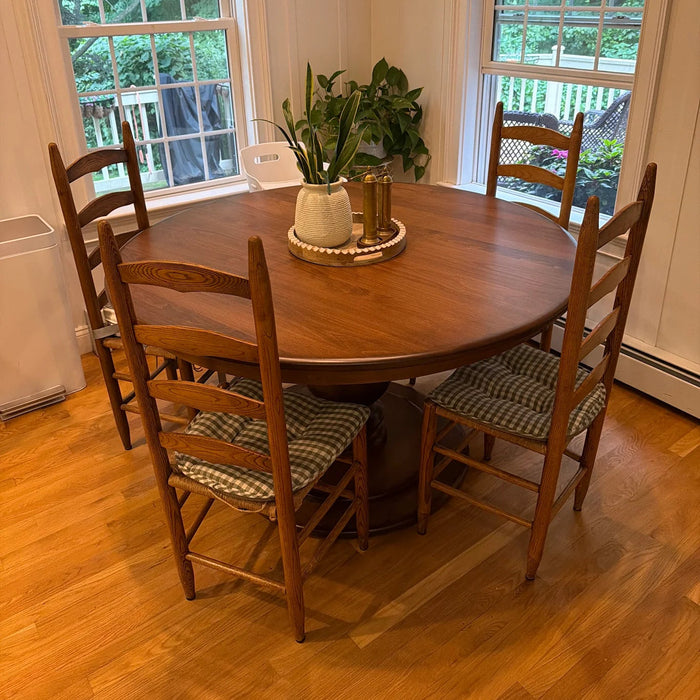 A round wooden dining table with four matching wooden chairs featuring striped cushions, set in a bright room with hardwood floors and windows looking out onto greenery.