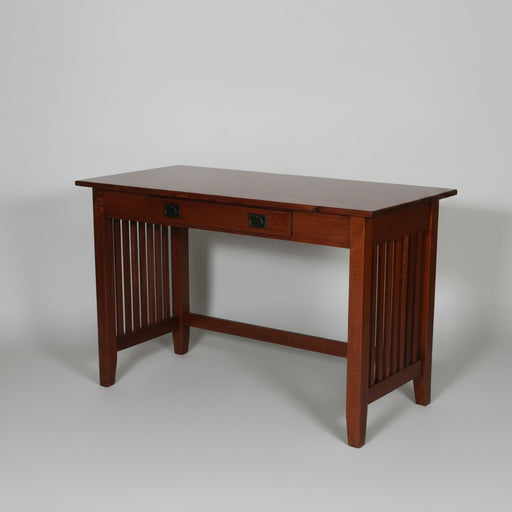 A dark reddish-brown wooden writing desk with a single drawer and decorative slatted side panels, shown with a green lamp, open book, and laptop on top against a plain light gray studio background.