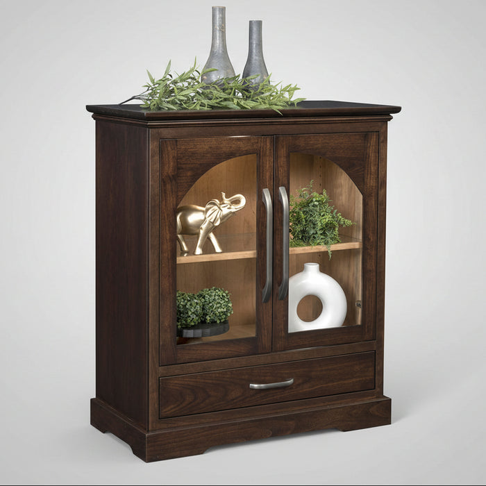 A dark brown wooden cabinet with two glass-front upper doors and a single drawer below, styled with decorative vases, greenery, and a small elephant figurine on a white background.
