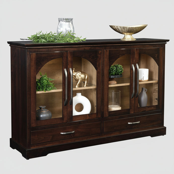 A dark brown wooden display cabinet with glass doors, shown against a solid white background. The cabinet features two upper doors with arched glass panels, two middle shelves with decorative items, two lower drawers with silver handles, and a dark wood top decorated with greenery and a gold bowl.