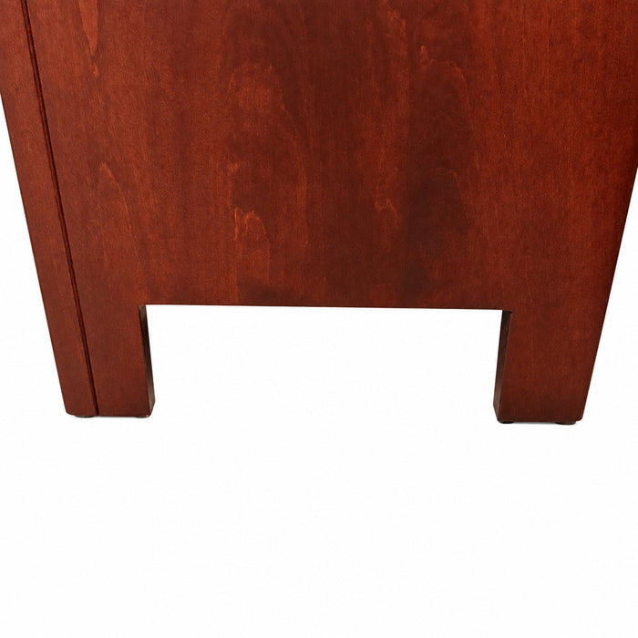 Close-up of the lower corner of a reddish-brown wooden furniture piece, likely a desk or cabinet, standing on a gray carpeted floor.