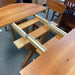 A wooden extendable dining table with attached benches and visible metal slide tracks, photographed in a furniture showroom with other chairs and tables around it.